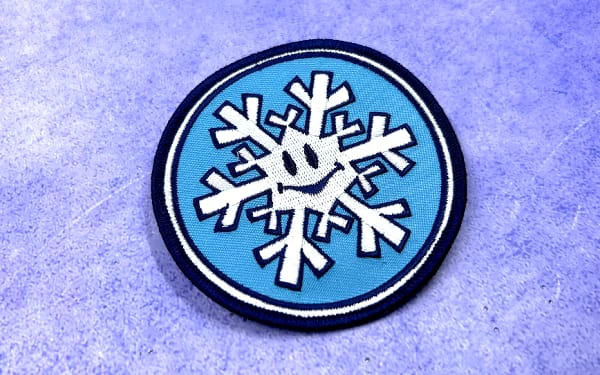 Round embroidered patch featuring a white snowflake with a smiling face on a light blue background. The patch has a dark blue border and is displayed on a textured purple surface.