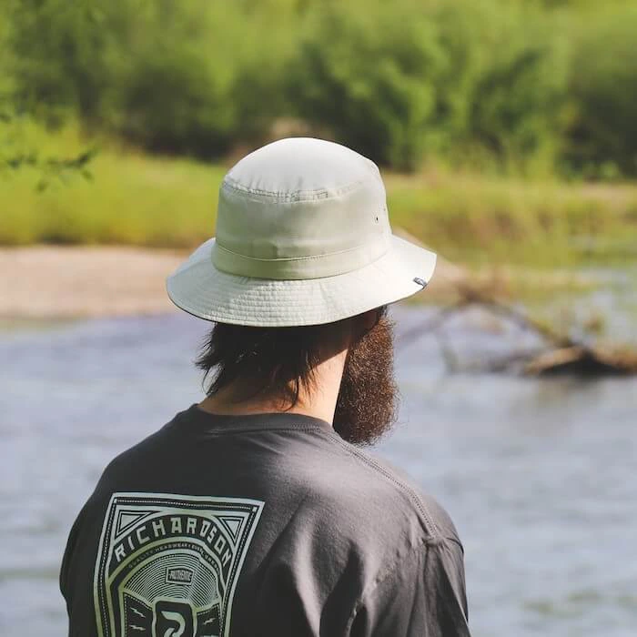 Man wearing a light-colored bucket hat while standing near a river, surrounded by lush greenery. He is wearing a dark Richardson-branded t-shirt, and the scene captures a relaxed outdoor setting.